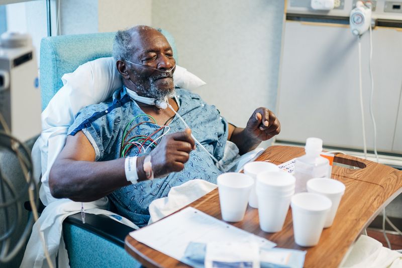 Patient with oxygen tube, eyes shut, hands lifted, cups on tray