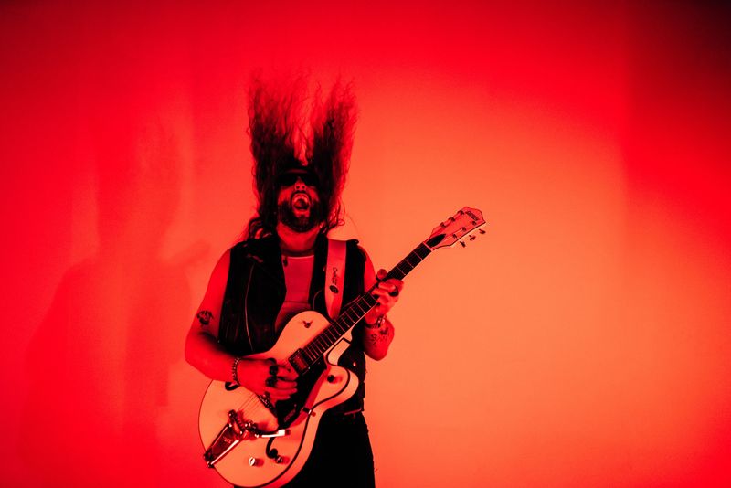 Hair blown straight up, shouting with guitar, flat red wall