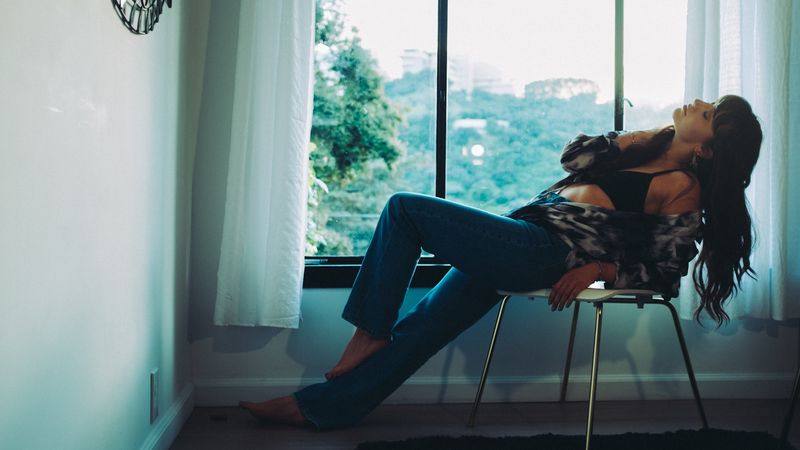A woman reclines in a chair at a window, a dark shape against green hills and white curtain