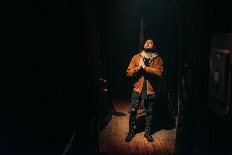 Hands pressed in prayer, eyes up, lone pool of backstage light