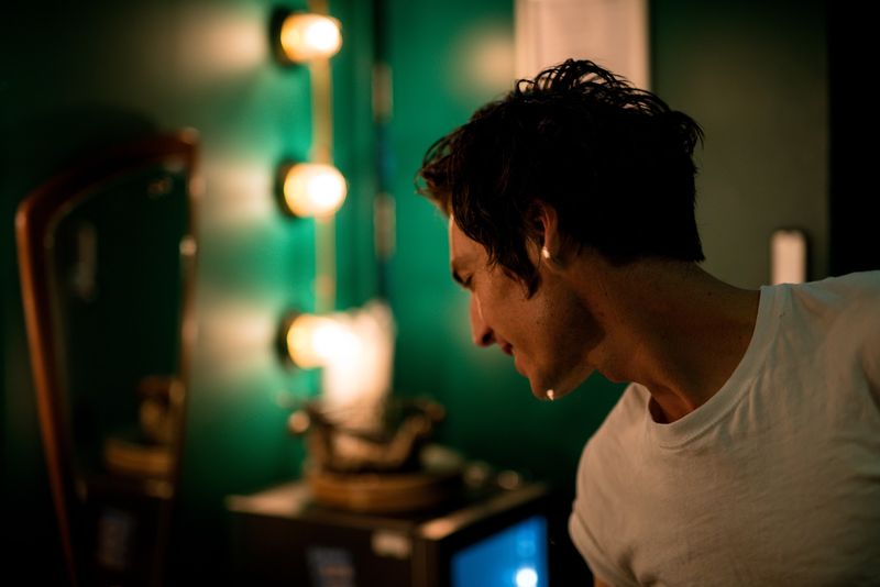 A man in a white tee looks down by dressing-room bulbs, green wall