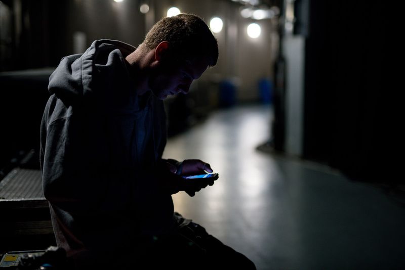 A man scrolls his phone in a dark corridor, bulbs soft behind