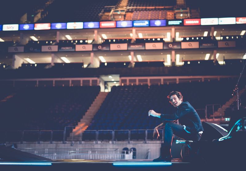 The singer crouches laughing at the stage edge of an empty arena