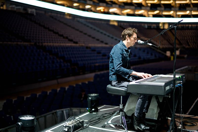 A pianist at soundcheck on a stage in an empty arena, rows of seats climbing