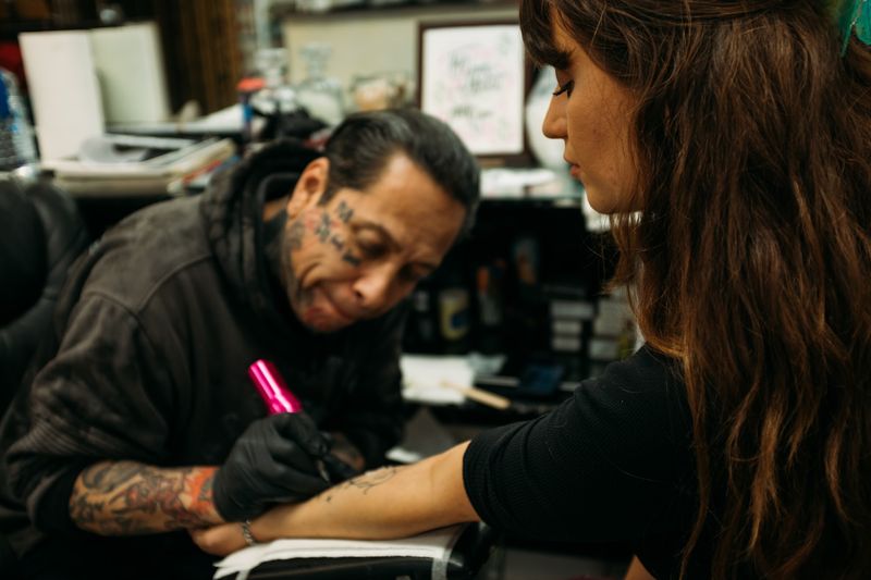 Face-tattooed artist marks a forearm with pink pen, intent