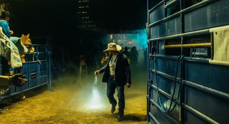 Cowboy walks rodeo chutes, flashlight beam on dirt, green haze