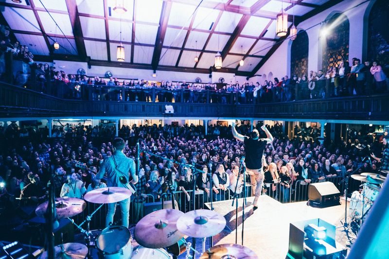 Packed hall and balcony from behind the drums, singer's arms up