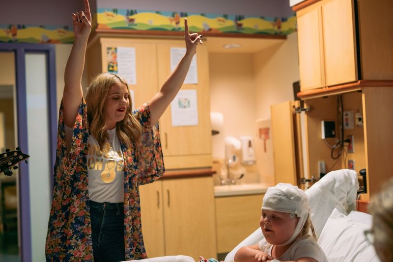 Singer's arms raised at a bedside, bandaged child beaming up
