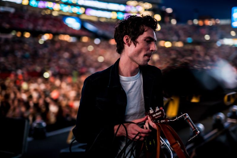 Musician side-stage with cables, stadium crowd glowing behind
