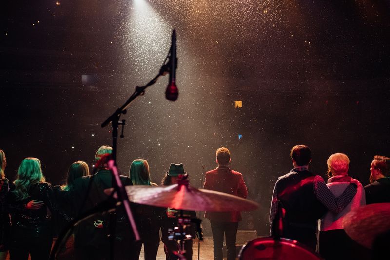 Cast arm in arm facing crowd as glitter falls in spotlight