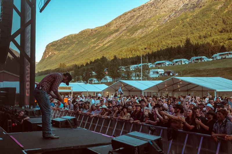 Artist leans to crowd, campers on sunlit Norwegian mountainside