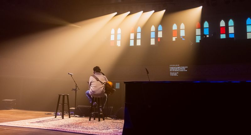 Lone guitarist on a rug, beams through stained-glass windows